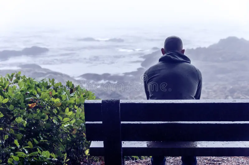 Homem de meia-idade sentado em um banco de praça, olhando pensativo para o horizonte.